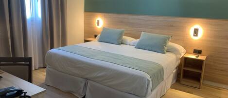 A neatly arranged hotel room with a large bed, bedside tables, a desk with a telephone, and a window with curtains.