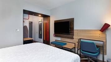A hotel room with a bed, a desk with a chair, a TV mounted on the wall, and a red lamp.