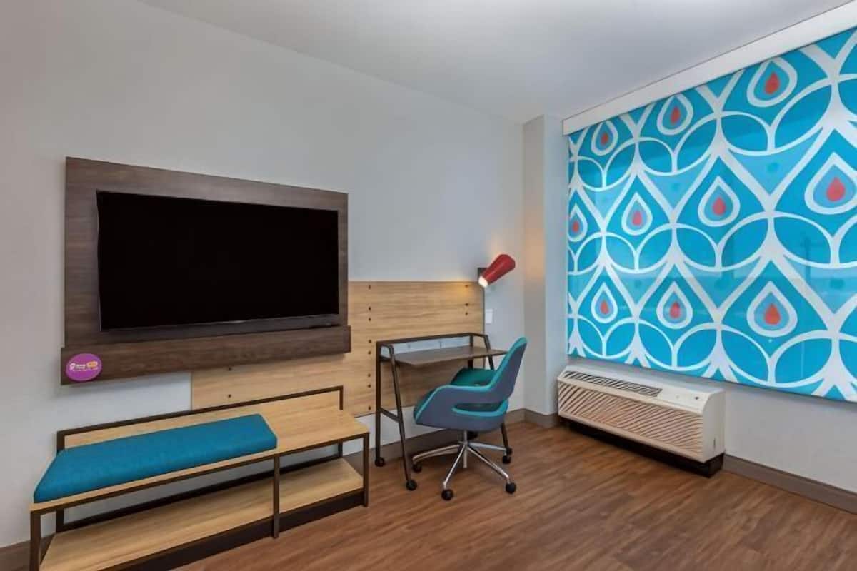 Een kamer met een grote flatscreen televisie aan de muur, een bureau met stoel, een bankje en een behang met patroon.