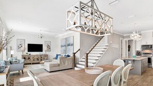 A spacious living room with a wooden dining table, white chairs, a staircase, and a kitchen area. - Coastal Luxury Cottage | Beach Access | Pet Friendly (Orange Beach)