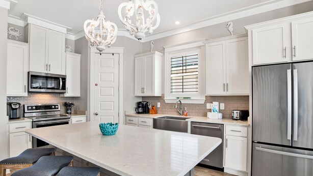 A modern kitchen with white cabinetry, a central island with bar stools, stainless steel appliances, and a chandelier. - Coastal Luxury Cottage | Beach Access | Pet Friendly (Orange Beach)