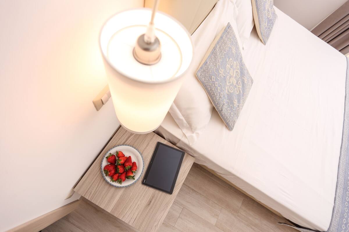 A neatly made bed with a pillow, a bedside lamp, a bowl of strawberries, and a tablet on a wooden bedside table.