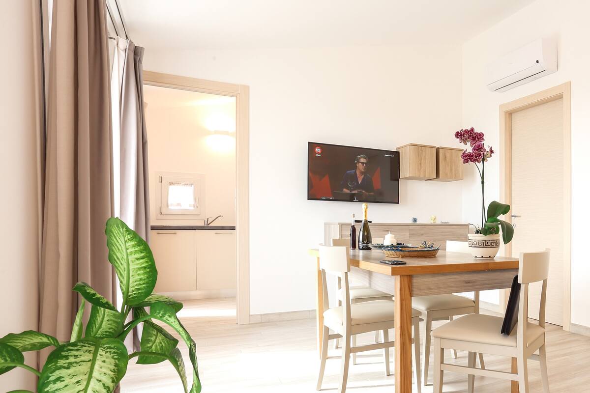 A modern living room with a dining area, a television mounted on the wall, and a potted plant.