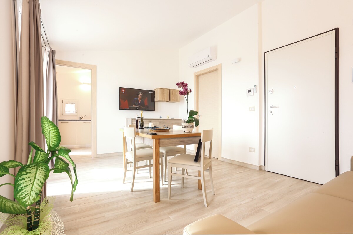 A modern living room with a dining area, a TV mounted on the wall, and a door leading to another room.