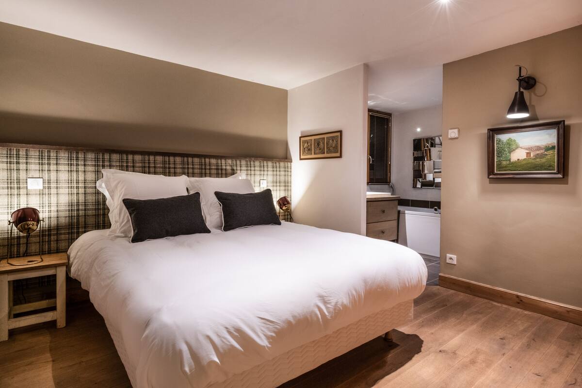 A bedroom with a large bed, bedside lamps, a small table, and a view of the kitchen.