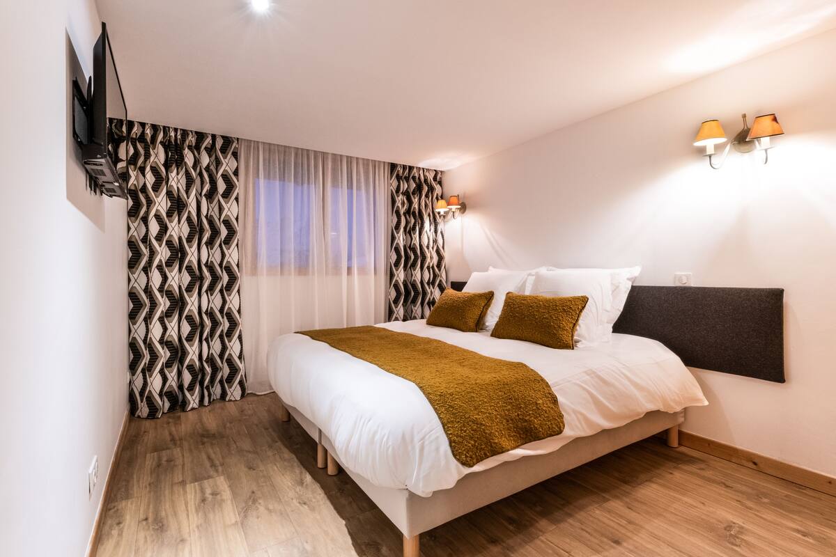 A bedroom with a bed, curtains, a flat-screen TV mounted on the wall, and a wall-mounted lamp.