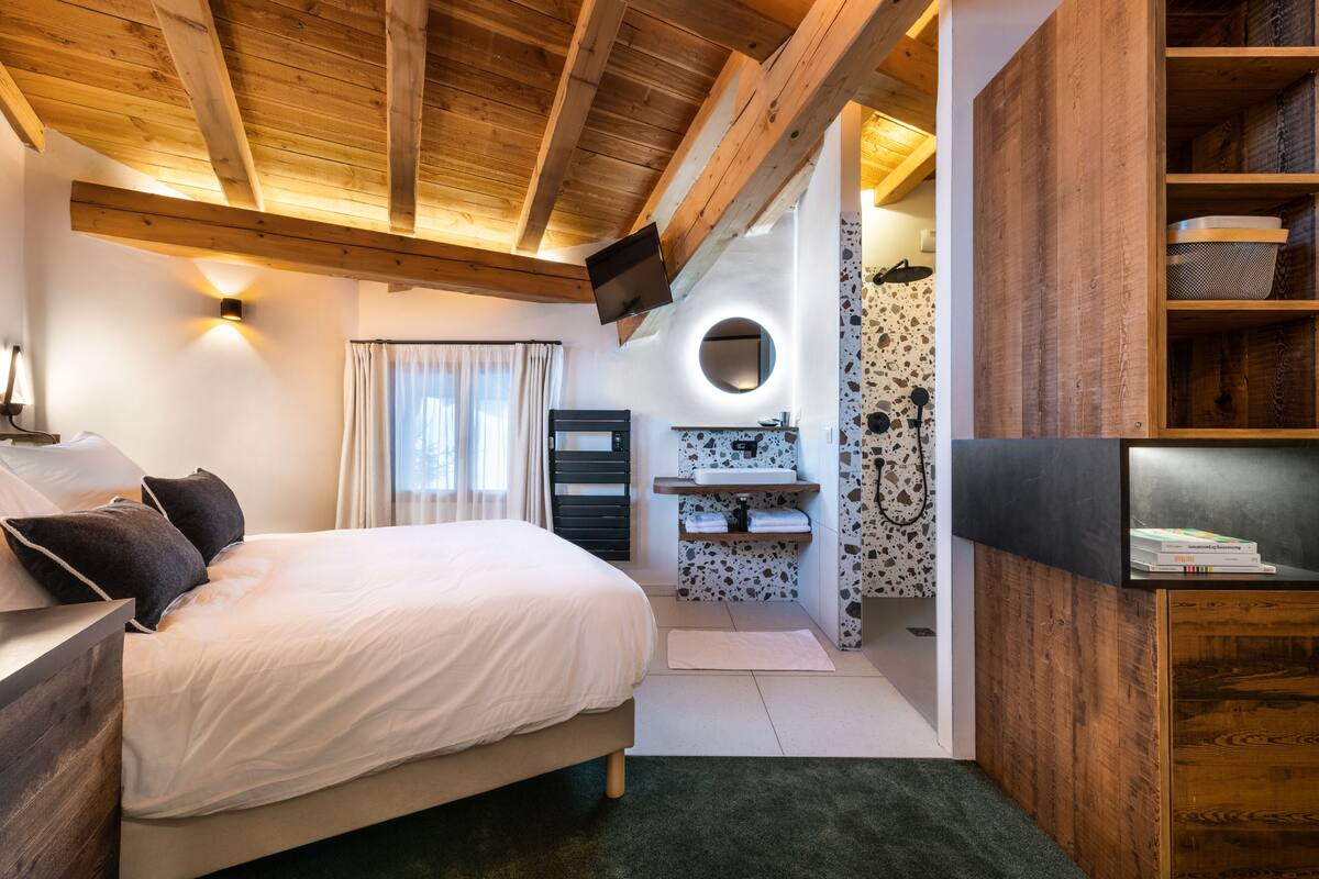 A bedroom with a bed, a TV, a bathroom with a sink and mirror, and wooden beams on the ceiling.