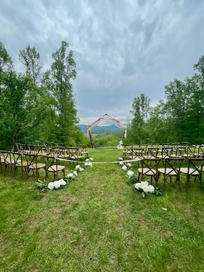 Un lieu de réception en plein air pour les mariages, avec une allée bordée de gazon, des chaises en bois et une arche décorée de fleurs.