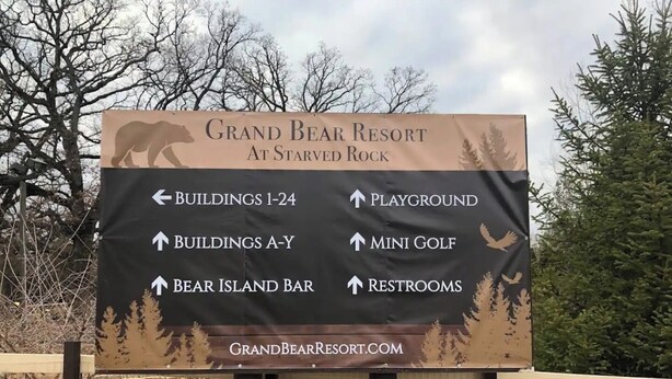 Un panneau de signalisation indiquant la direction du Grand Bear Resort, situé à Starved Rock, avec des flèches pointant vers les bâtiments, l’aire de jeux, le mini-golf et les toilettes.