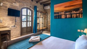 A hotel room with a stone wall, a bed with a blue bedspread, a mounted TV, and a painting of a sunset over a harbor.