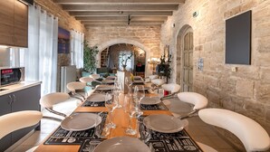 A dining area with a long table set for six, surrounded by white chairs, in a room with stone walls and wooden beams.