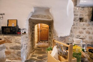 Cozinha com paredes de pedra e uma porta de madeira que dá para outro cômodo, um micro-ondas e uma tábua de corte de madeira no balcão.