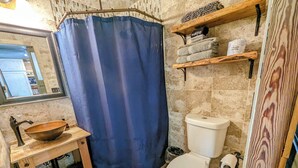 A bathroom with a wooden vanity, a large bowl sink, a toilet, a shower curtain, and shelves with towels and toiletries.
