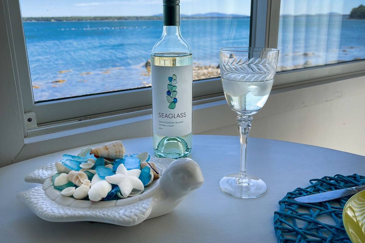 A bottle of Seaglass Sauvignon Blanc, a glass of water, and a decorative plate with seashells and starfish on a table by a window with a view of the sea.