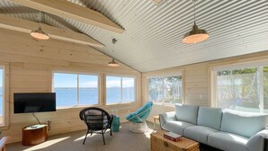 A spacious living room with a large window overlooking the sea, a comfortable sofa, a TV, and a wicker coffee table. - Acadia Bay Cottages (Surry)