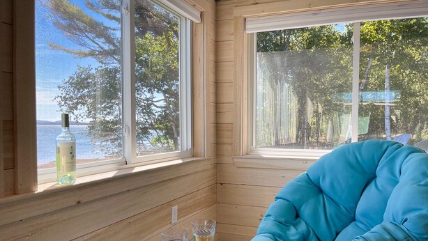 A room with wooden walls and large windows overlooking a body of water and trees. There is a blue cushioned chair, a small table with two glasses of white wine and a plate of food. - Acadia Bay Cottages (Surry)