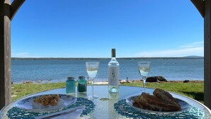 A table with a bottle of wine, two glasses, and plates of food by a lakeside view. - Acadia Bay Cottages (Surry)