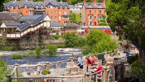 A riverside restaurant with outdoor seating overlooking a flowing river, surrounded by historic buildings. - Bridge Hideaway (Llangollen)