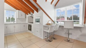 Una cocina con armarios blancos, una zona de comedor con dos taburetes de bar, una ventana con persianas y un techo con vigas de madera.