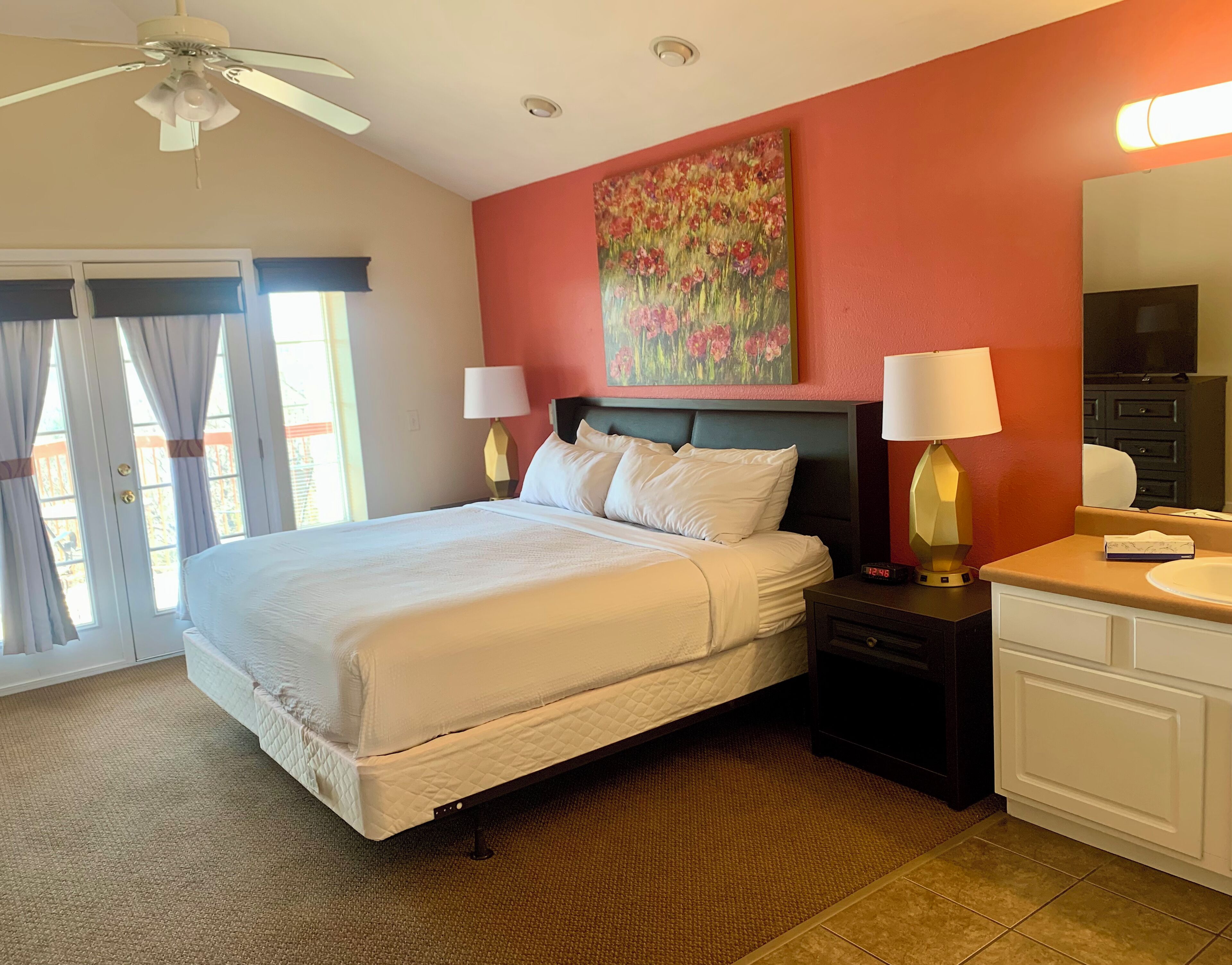 A hotel room with a bed, two bedside lamps, a painting on the wall, a ceiling fan, and a bathroom with a sink and mirror.