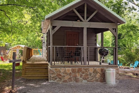 A wooden cabin with a porch, stone foundation, and a grill outside.