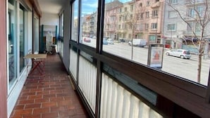 Un corridoio stretto con pavimento in mattoni, ampie finestre e vista su una strada con auto parcheggiate e palazzi.