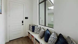 Ein Flur mit einer weißen Tür, einer Bank mit gemusterten Kissen und einem Fenster mit Blick auf ein Haus.