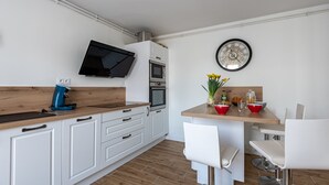 Cozinha moderna com armários brancos, bancada de madeira, forno embutido, micro-ondas, cafeteira e um espaço de jantar com mesa de madeira e cadeiras brancas.