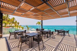 A covered outdoor seating area with tables and chairs, overlooking a pool and the ocean.