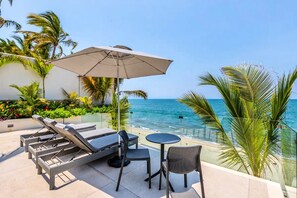Een zonnig terras buiten met lounge fauteuils, een tafel en een parasol met uitzicht op een strand.
