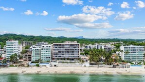 A coastal cityscape with modern buildings, a beach, and a mountainous backdrop. - CDM 205: Beachfront 2BR w Pool, Gym & Ocean Views (Bucerías)