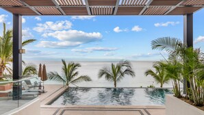 A pool with a view of the sea, surrounded by palm trees and a covered area with chairs and umbrellas. - CDM 205: Beachfront 2BR w Pool, Gym & Ocean Views (Bucerías)