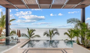 A pool with a view of the sea, surrounded by palm trees and a covered area with chairs and umbrellas.