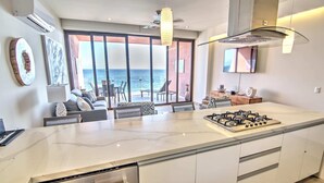 A modern kitchen with a large island, a gas stove, and a view of the ocean through large windows. - CDM 205: Beachfront 2BR w Pool, Gym & Ocean Views (Bucerías)