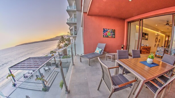 A balcony with a view of the ocean, a dining table with chairs, and a lounge chair. - CDM 205: Beachfront 2BR w Pool, Gym & Ocean Views (Bucerías)