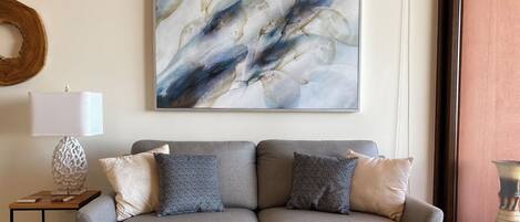 Una sala de estar moderna con un sofá gris, dos almohadones estampados, un cuadro abstracto colgado en la pared, una mesita de noche con una lámpara y un anillo decorativo de madera.