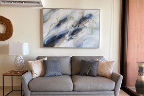 Een moderne woonkamer met een grijze bank, twee kussens met een patroon, een wanddecoratie in abstractie, een bijzettafeltje met een lamp, en een decoratieve houten ring.