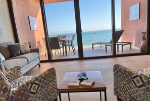 A living room with a sofa, two patterned armchairs, a coffee table with books and a remote control, and a large sliding glass door leading to a balcony with a view of the sea. - CDM 205: Beachfront 2BR w Pool, Gym & Ocean Views (Bucerías)