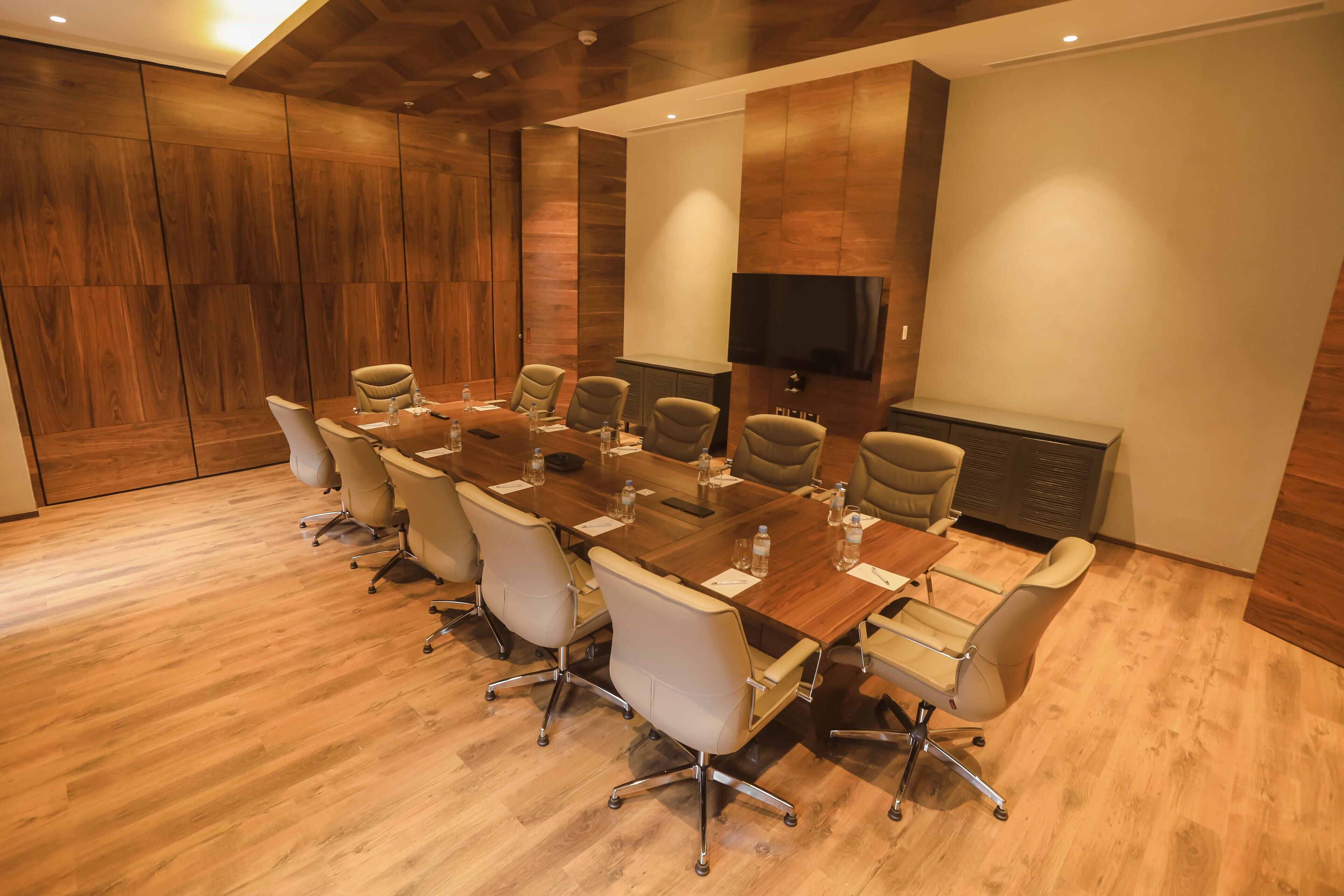 A conference room with a long table, multiple chairs, a flat-screen TV, and wooden paneling on the walls.