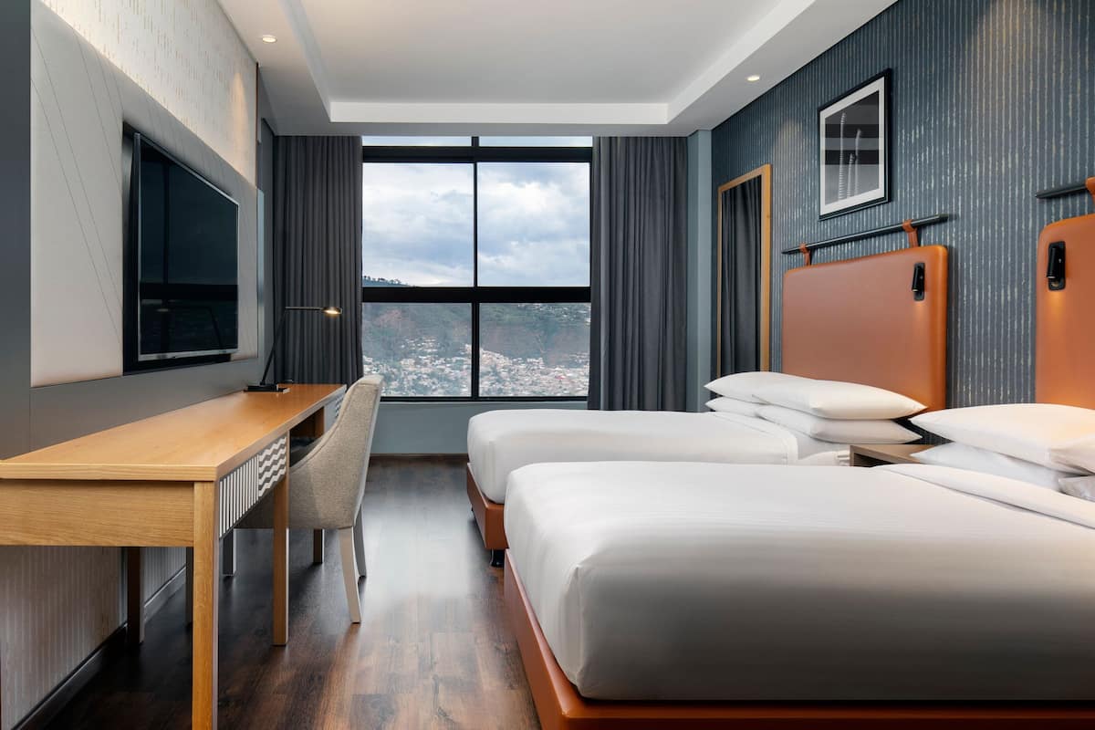 Habitación de hotel moderna con un ventanal grande, un escritorio, dos camas y un televisor montado.