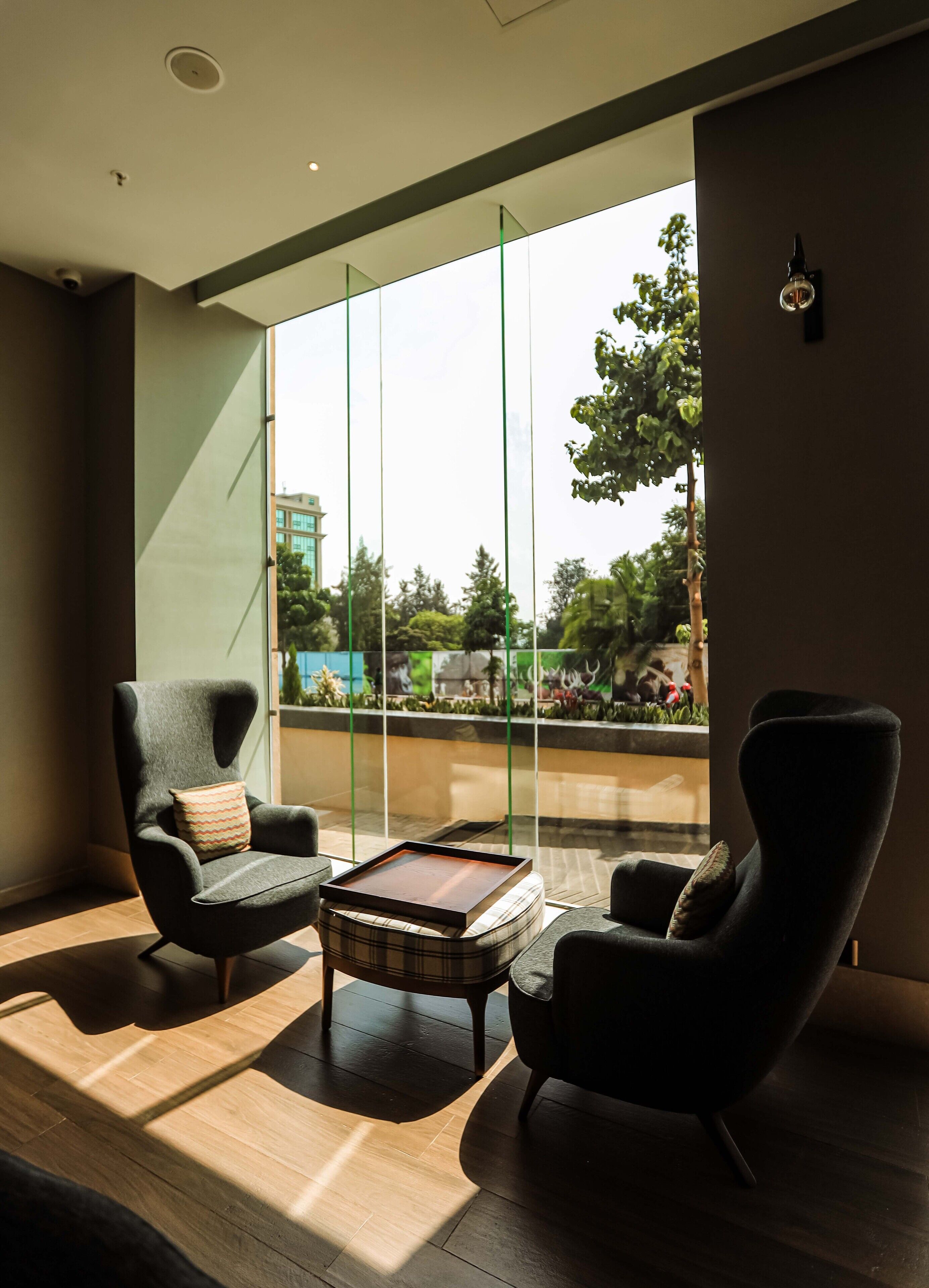 A modern lounge area with two armchairs, a wooden coffee table, and a large window offering a view of greenery and buildings.