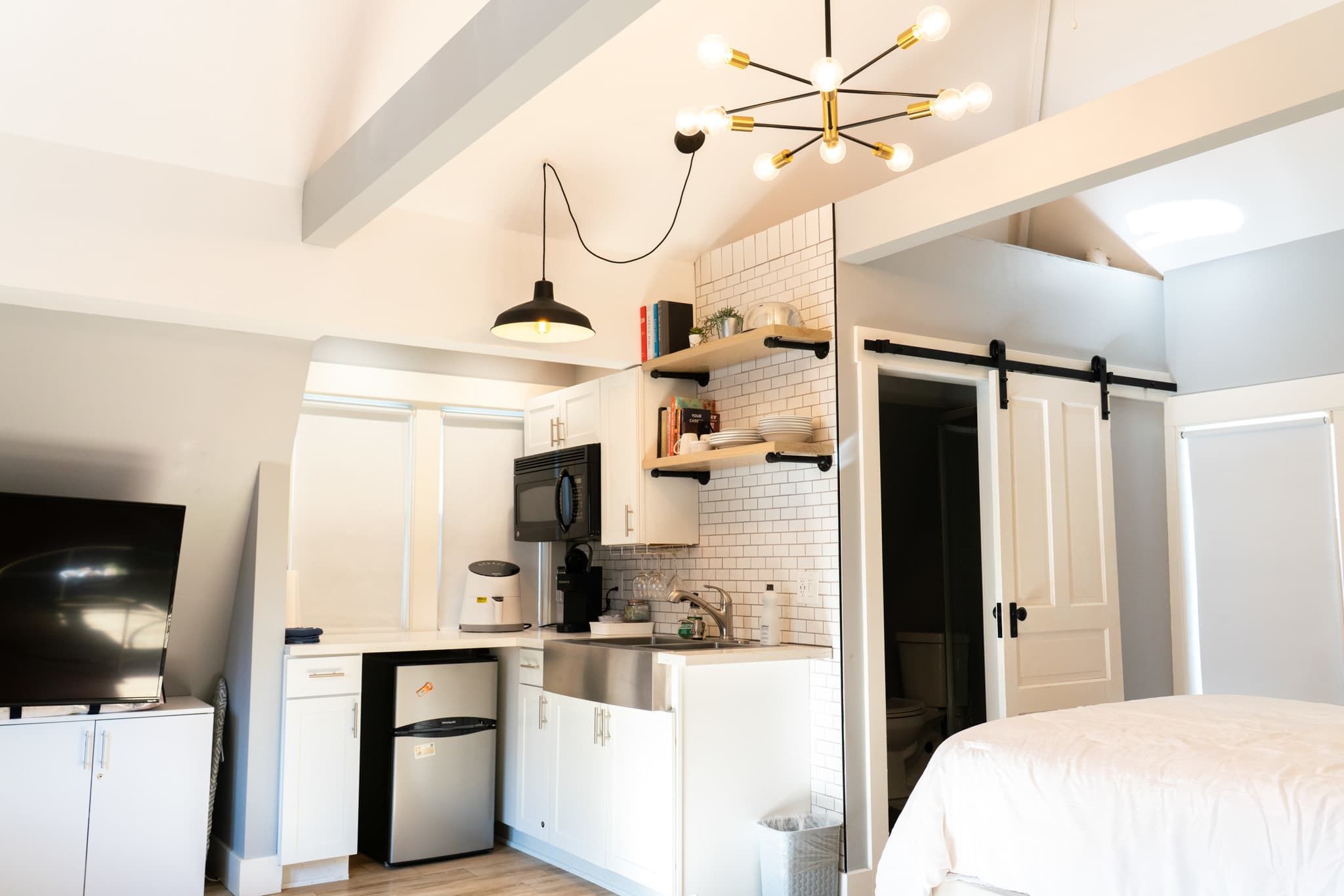 A compact kitchen with white cabinetry, a microwave, a coffee maker, and a sink. There is a sliding door leading to a bathroom.
