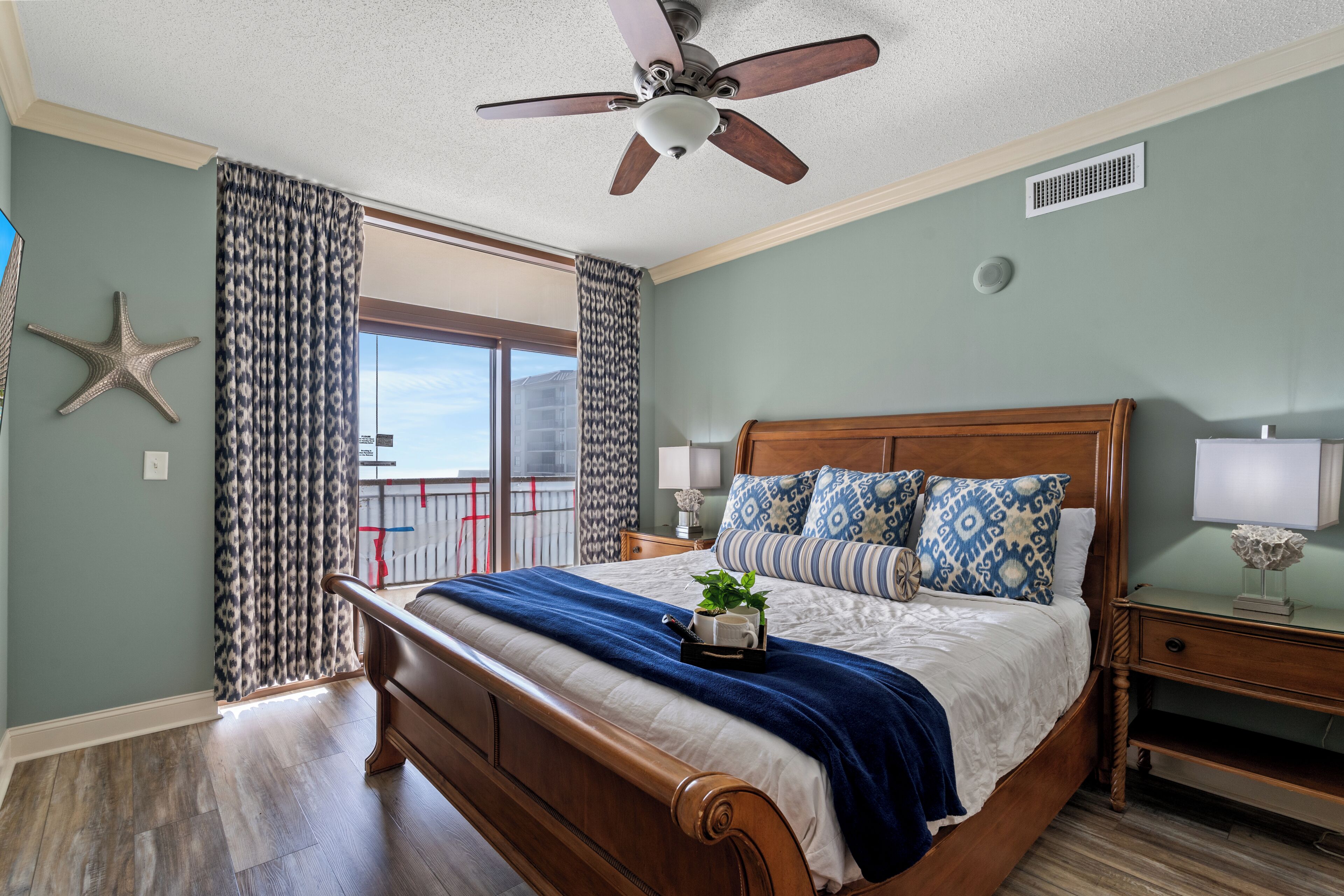 A bedroom with a wooden bed, a ceiling fan, a nightstand with a lamp, and a view of the outdoors.