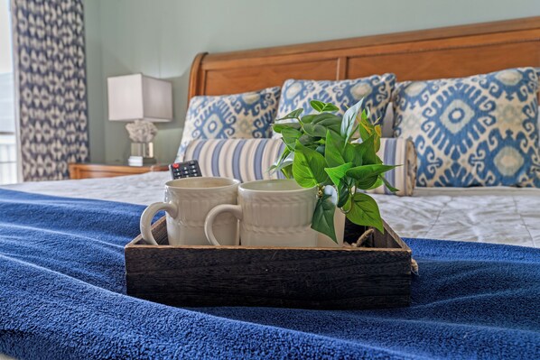 Una cama con un edredón azul, una bandeja de madera con dos tazas blancas, una planta en maceta y almohadas estampadas.