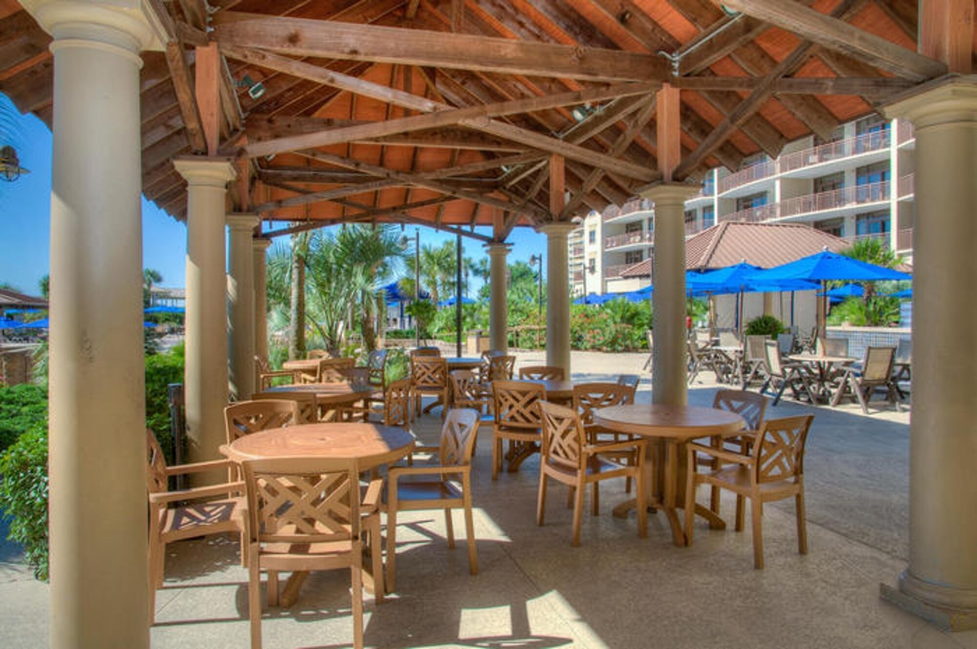 Un espacio al aire libre con un patio, mesas y sillas de madera, rodeado de palmeras y una piscina al fondo.