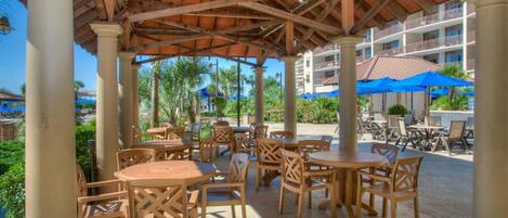 Un espacio al aire libre con un patio, mesas y sillas de madera, rodeado de palmeras y una piscina al fondo.