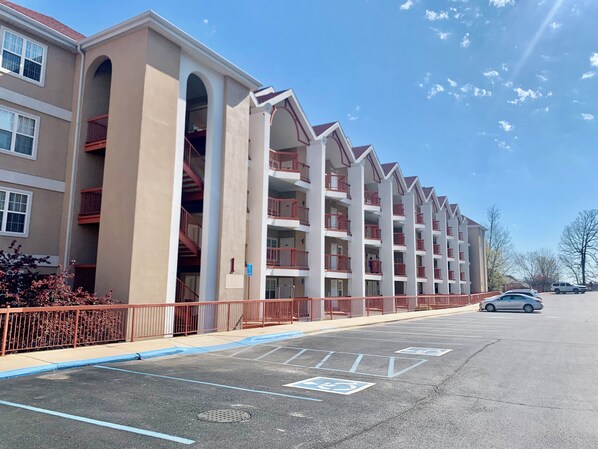 Un hotel de varios pisos con fachada color crema, barandillas rojas y un estacionamiento con espacios designados para personas con discapacidad.