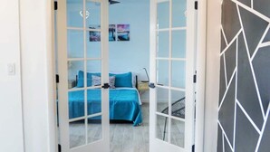Quarto com uma cama, uma escrivaninha e uma cadeira. Há uma porta que dá para outro espaço.