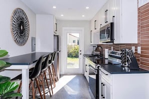 A compact kitchen with white cabinets, a black countertop, and a microwave. There are bar stools and a large decorative wall piece.