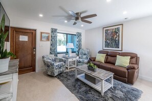 Ein Wohnzimmer mit Deckenventilator, einem braunen Ledersofa, gemusterten Sessel, einem Couchtisch und einem großen Fenster mit Vorhängen.
